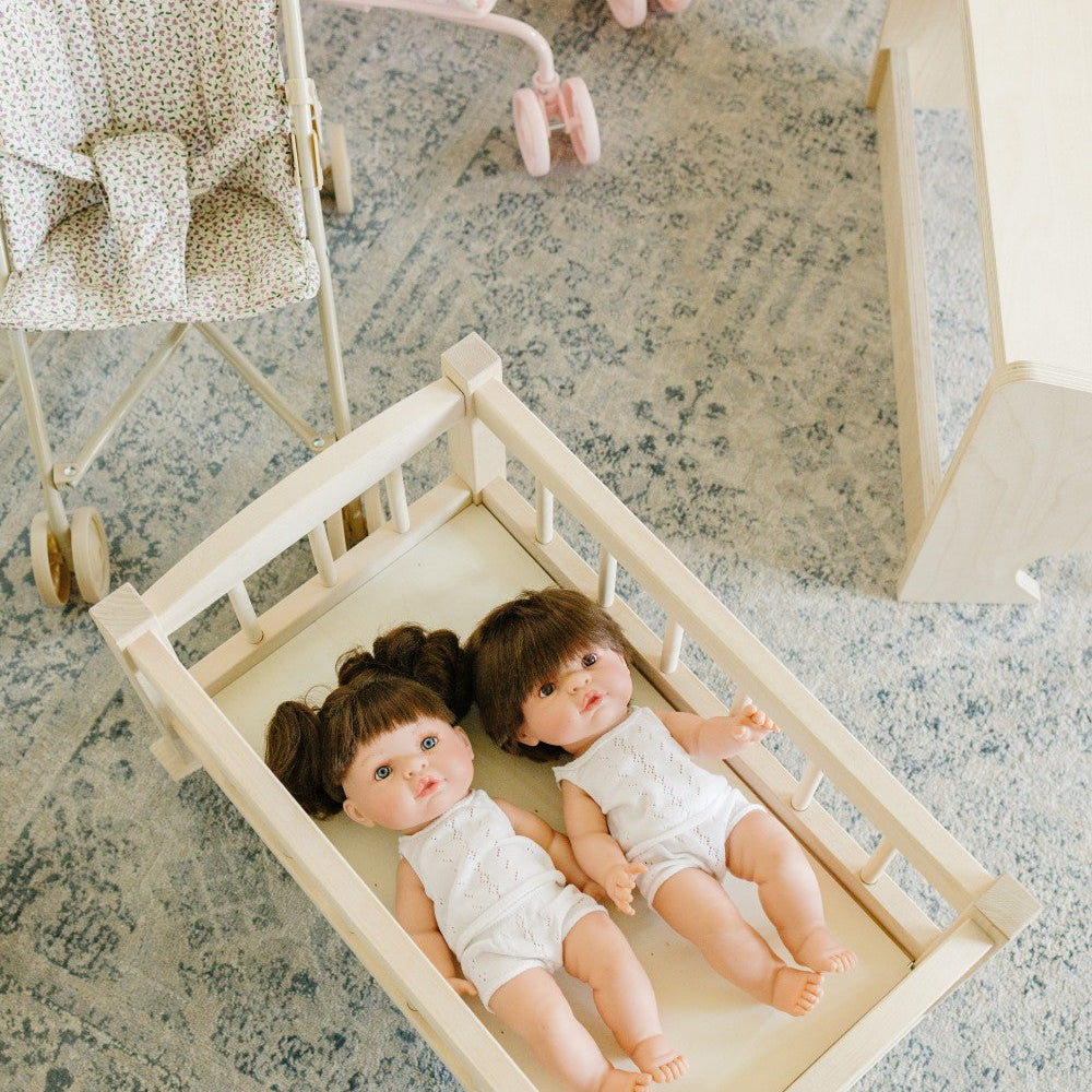 Two baby dolls lying on a cream fabric mattress inside a light wood bassinet, placed on a blue-gray patterned rug in a nursery setting with a stroller and changing table visible in the background.