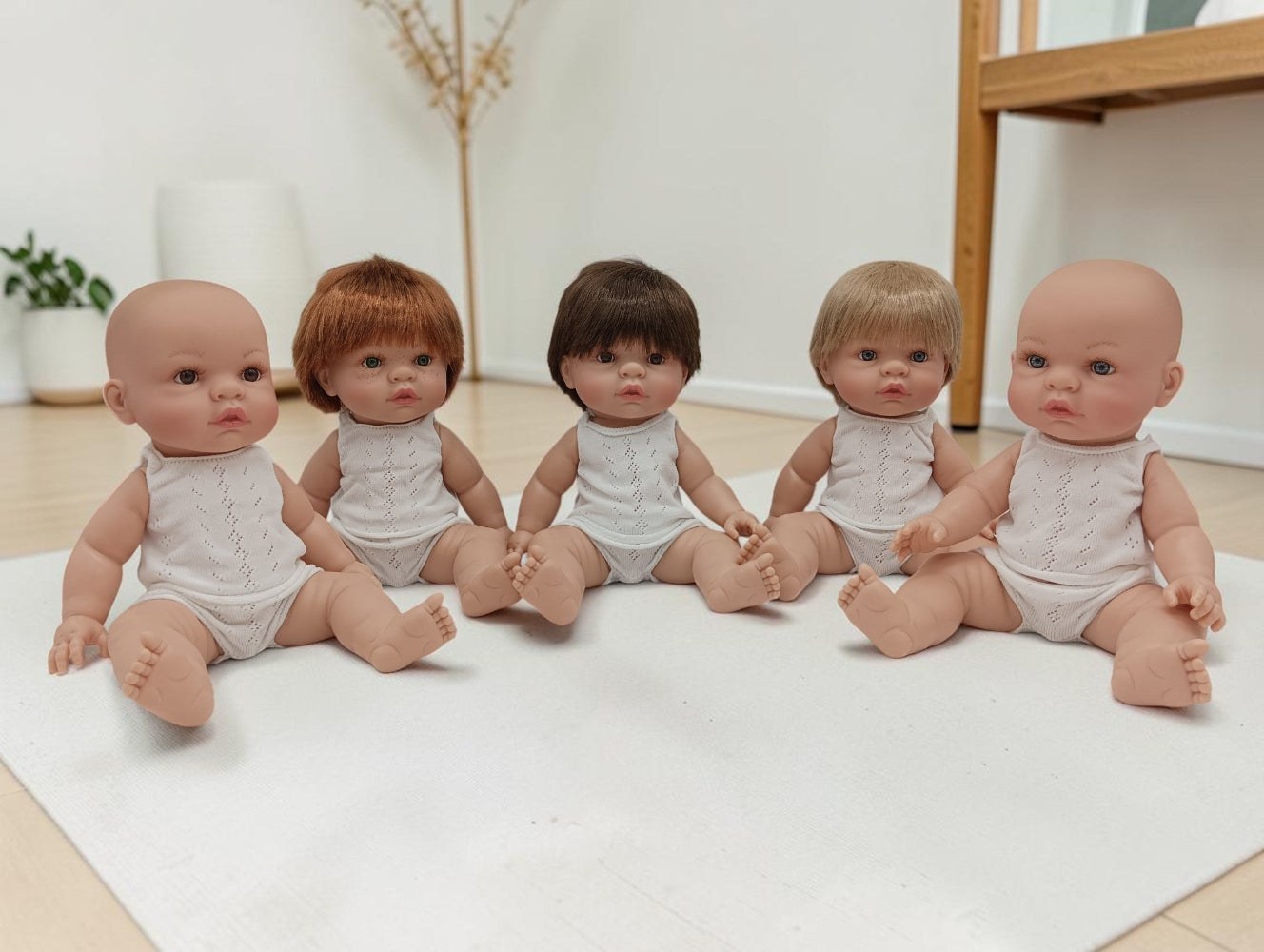 Five baby dolls with different hair colors sitting on a white mat.