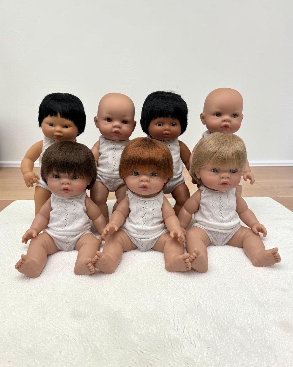 Sevenrealistic baby dolls with various hair colors and skin tones, wearing white outfits, arranged on a white textured rug against a plain wall.
