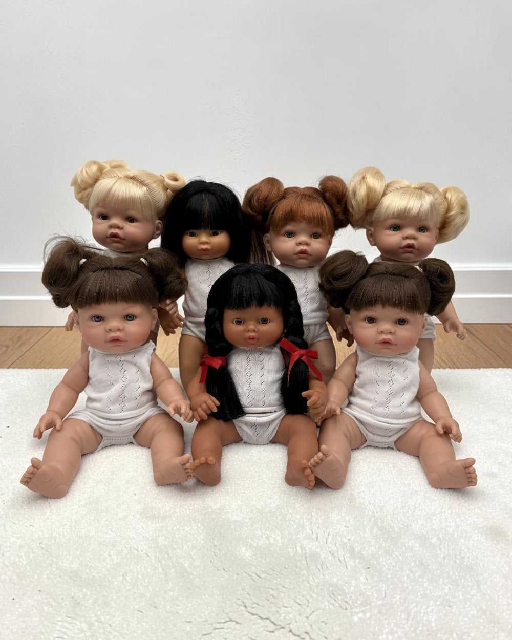 Seven realistic baby dolls with diverse hair colors and styles, wearing white outfits, arranged together on a white textured rug against a plain white wall.