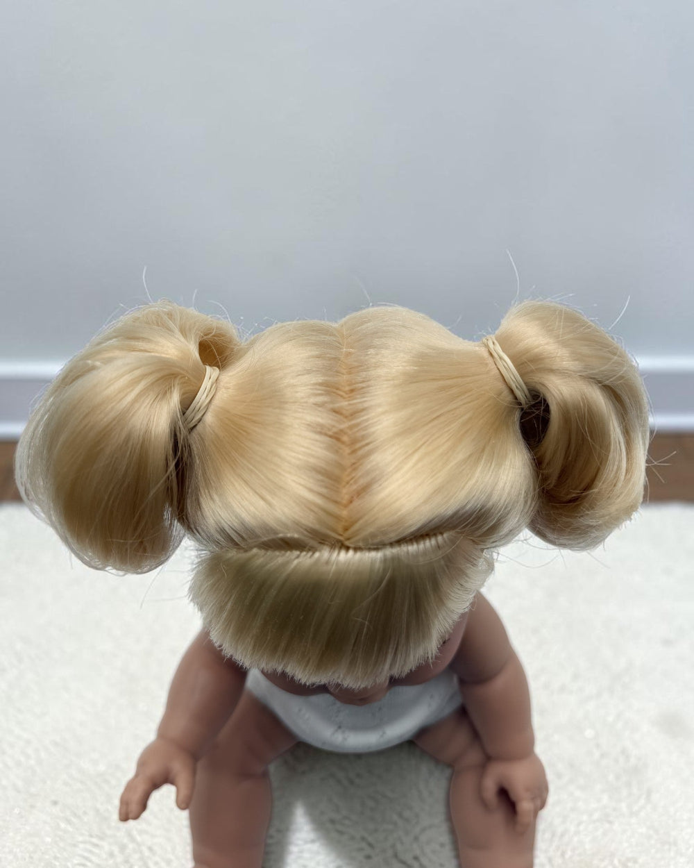 A doll viewed from behind, wearing a blonde hair accessory with two pigtails and a center part, seated on a white textured surface against a plain light gray wall.