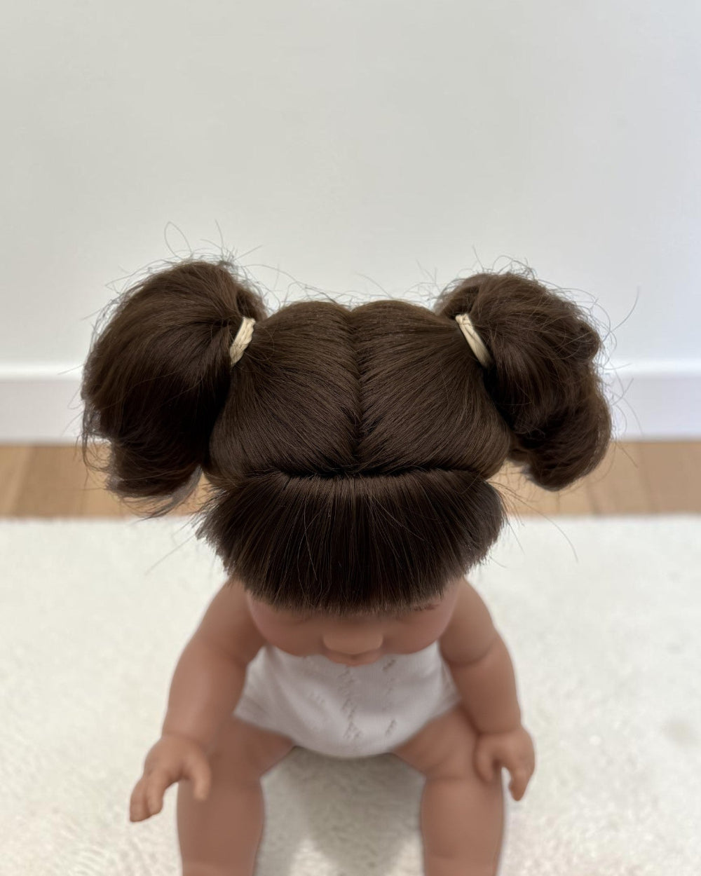 A doll with dark brown hair styled in two buns with small cream hair ties, wearing a white diaper, sitting on a white textured rug.
