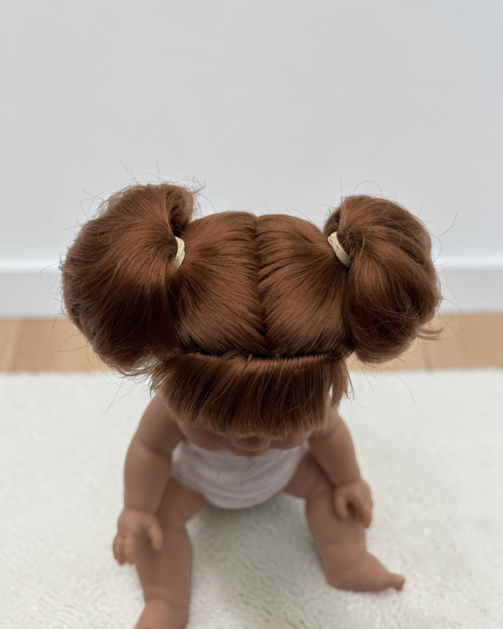 A close-up shot of a doll from behind, showing its detailed auburn hair styled in two buns with small cream elastic bands, wearing a white top, sitting on a white textured rug.