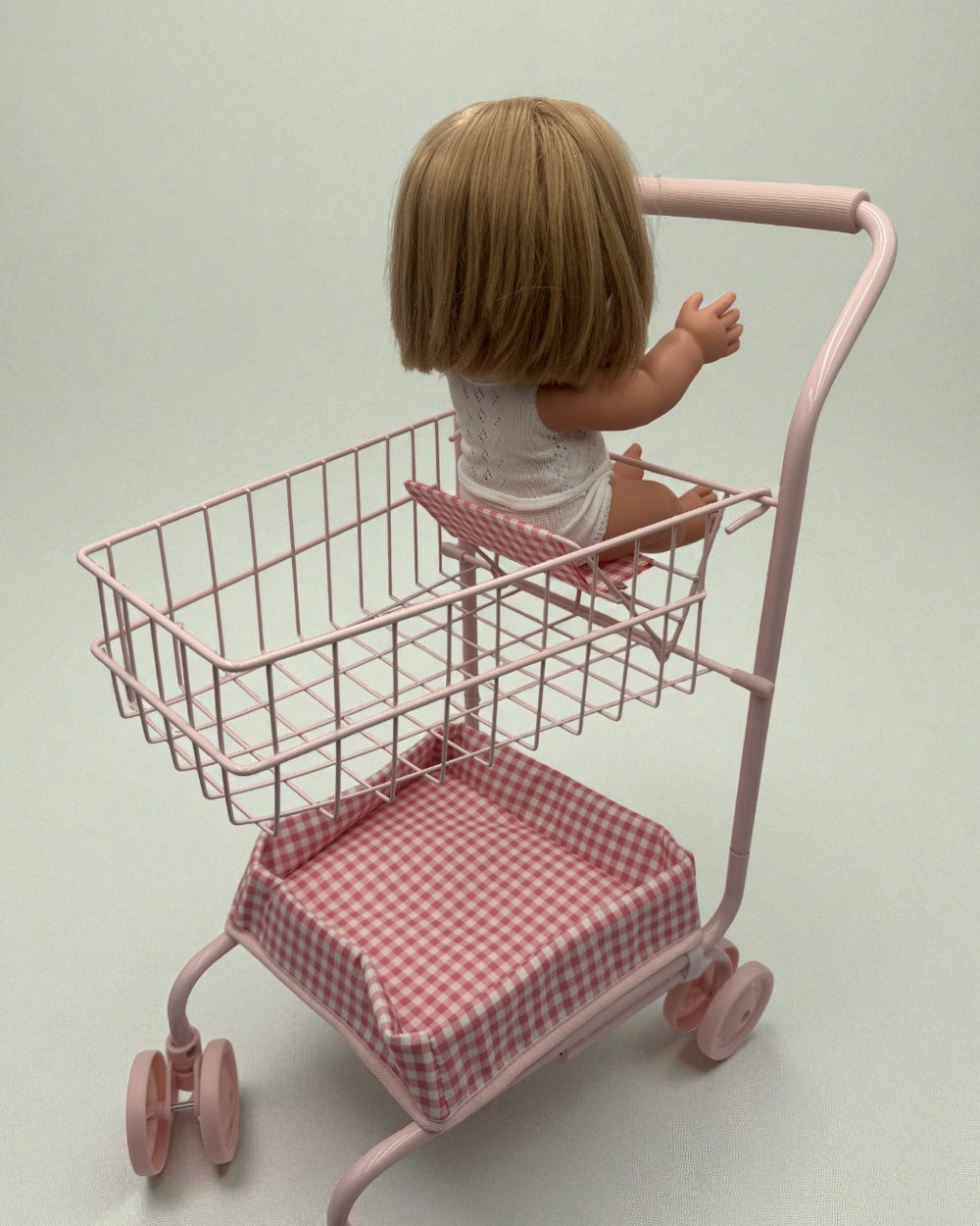 Doll sitting in a pink toy shopping cart with a checkered seat on a plain background
