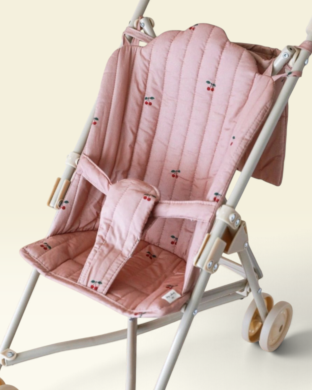 A close-up of a baby stroller seat cover in dusty pink with a small cherry print, shown fitted on a stroller frame against a light cream background.
