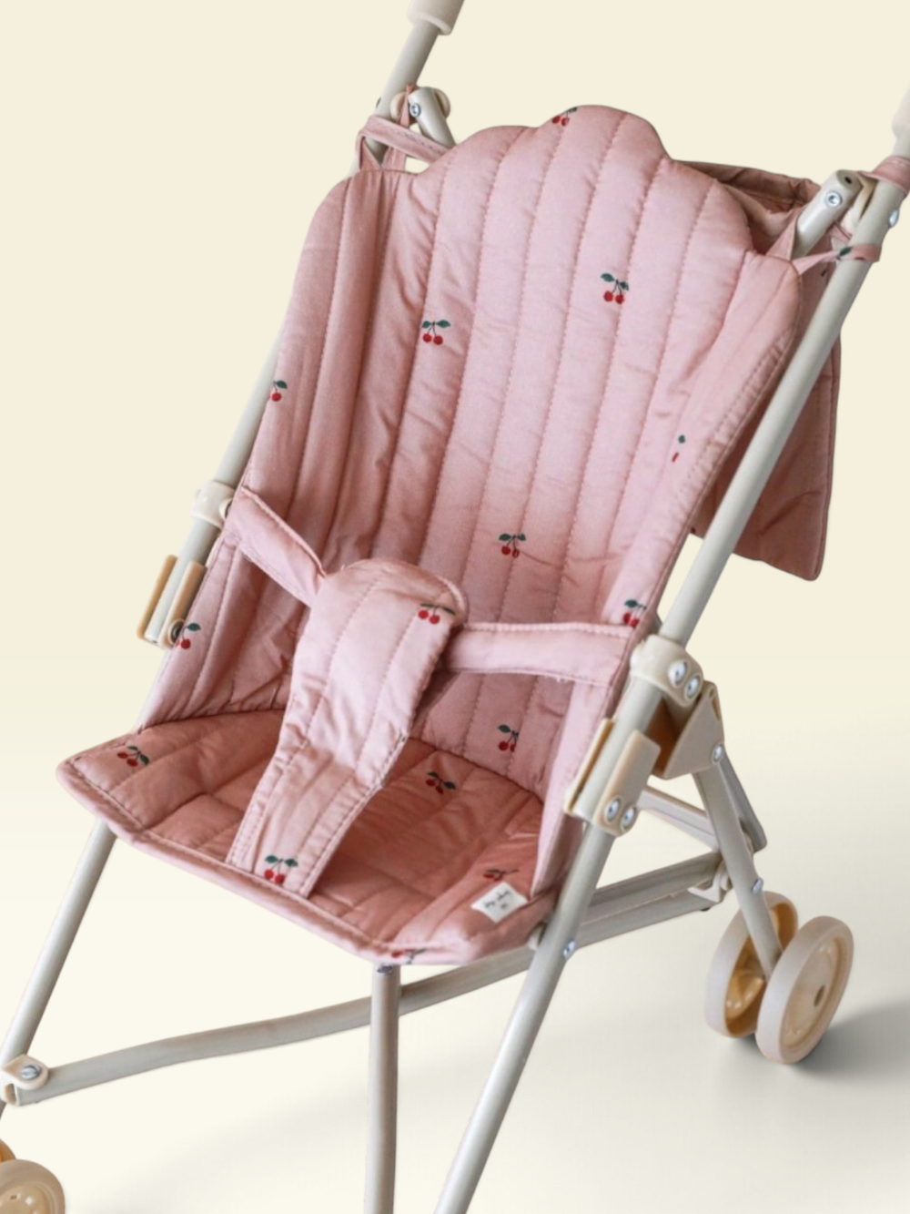 A close-up of a baby stroller seat cover in dusty pink with a small cherry print, shown fitted on a stroller frame against a light cream background.
