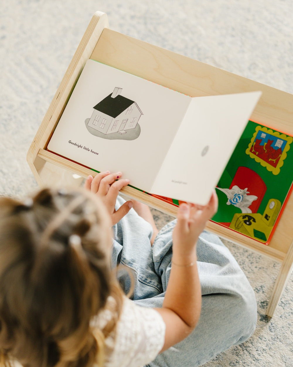 A child sitting on a patterned carpet reads from an open wooden children's book stand. The book shows a simple house illustration, and colorful children's books are tucked into the side of the stand.