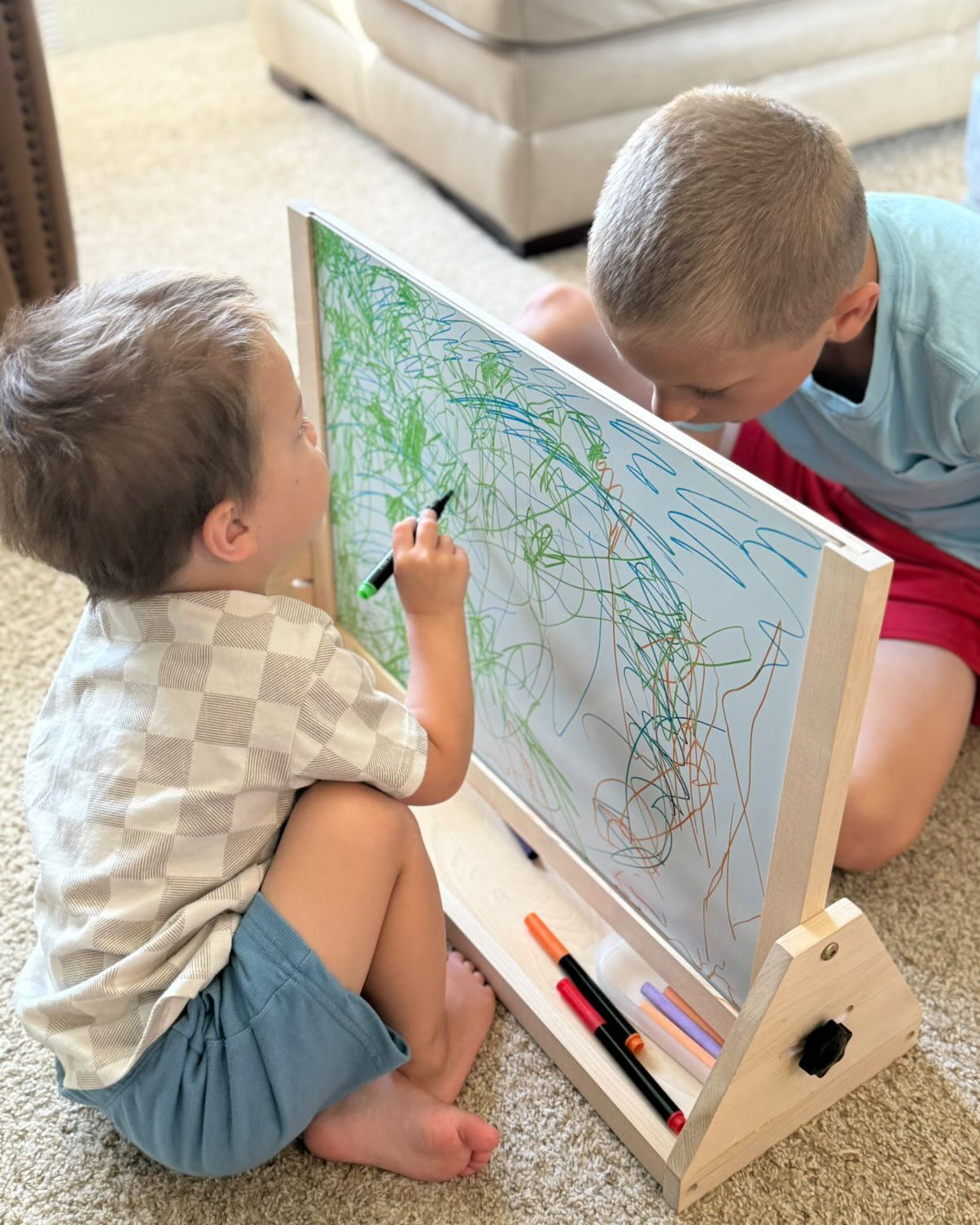 Two children drawing on a whiteboard with markers in a living room setting.