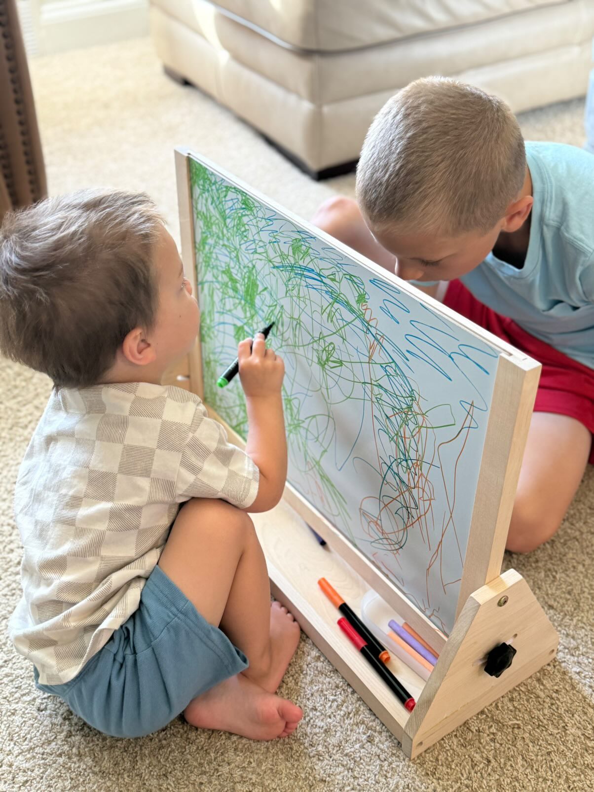 Two children drawing on a whiteboard with markers in a living room setting.