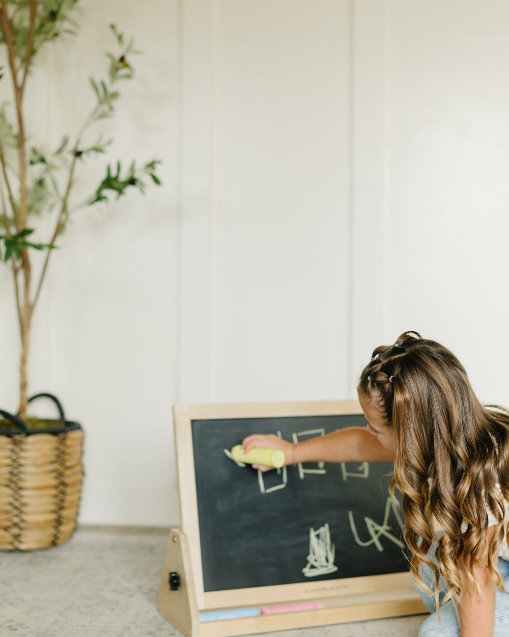 A young girl with long wavy brown hair kneels on a gray carpet, writing on a small wooden children's chalkboard. The chalkboard has a black slate face, a natural wood frame, and a light wood base with chalk sticks.