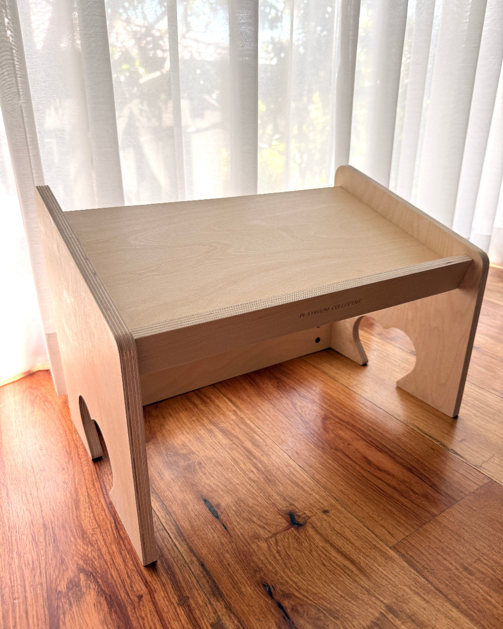 A light natural wood folding desk or stand with a slanted top, shown on a hardwood floor in front of sheer white curtains.