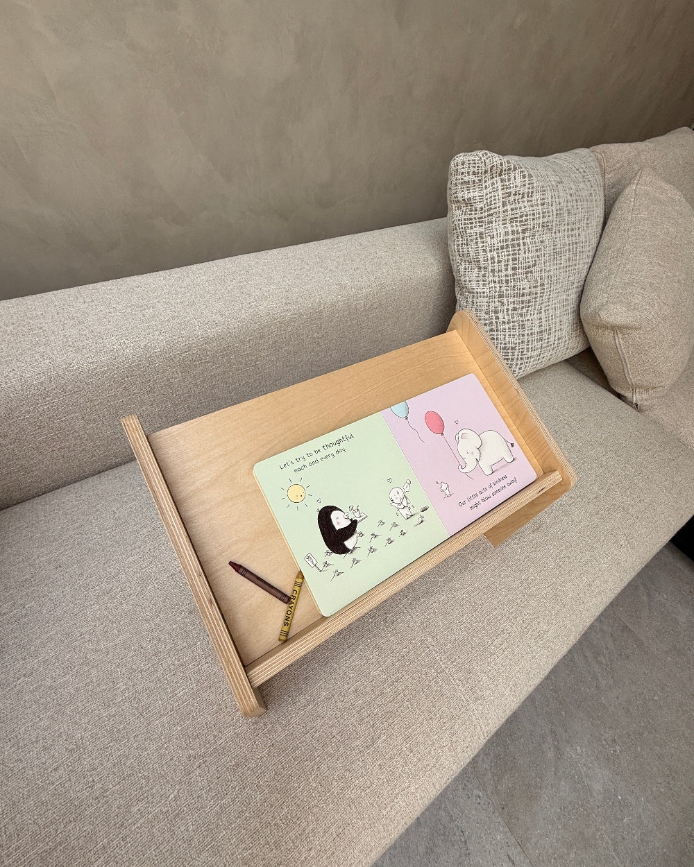 A light wood lap desk tray resting on a gray fabric sofa, holding a pastel cartoon card and a pencil.