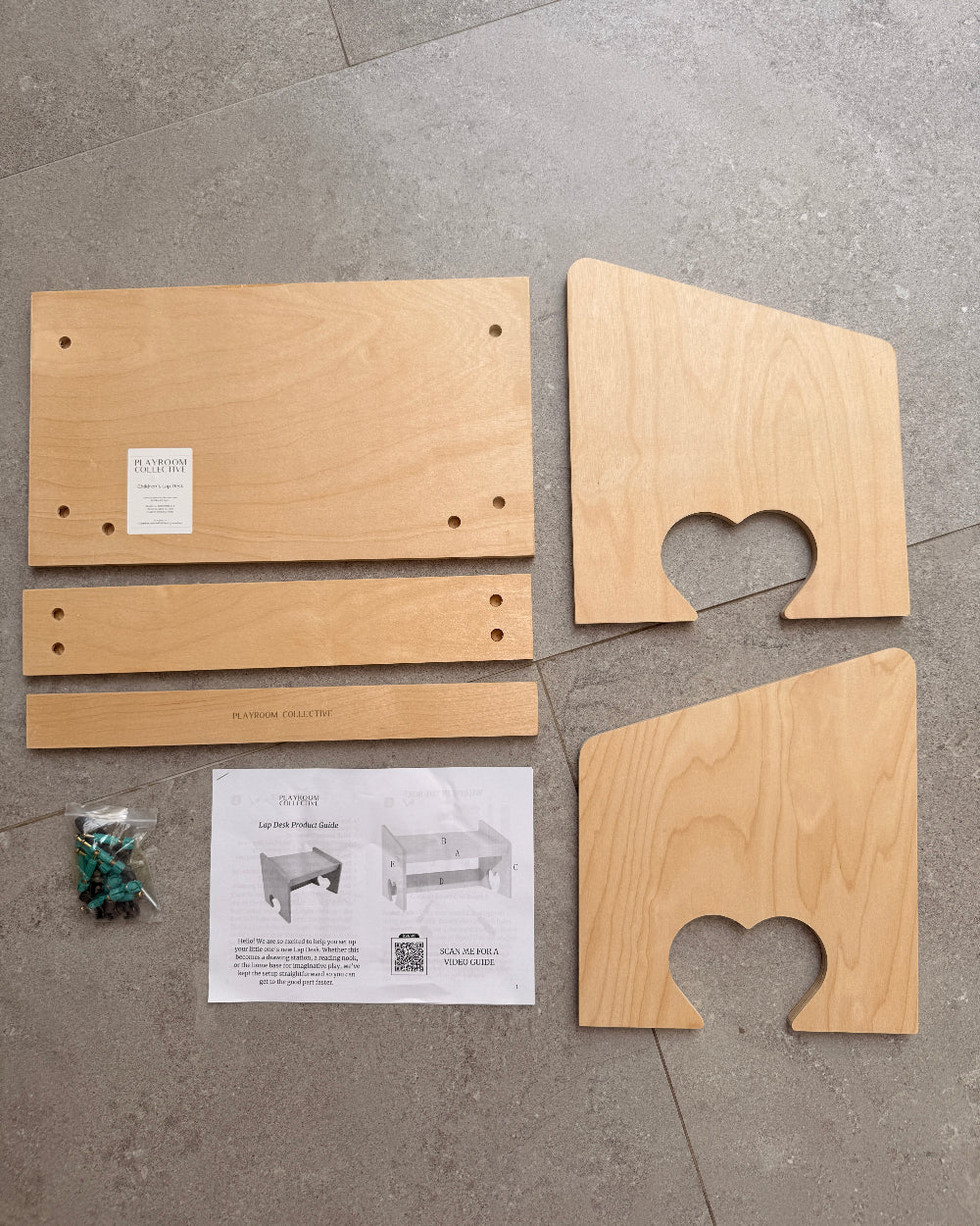 Wooden furniture assembly kit parts laid out on a gray tiled floor, including two curved top panels, two rectangular side panels, a long narrow piece, a printed instruction sheet, and a small bag of screws and hardware.