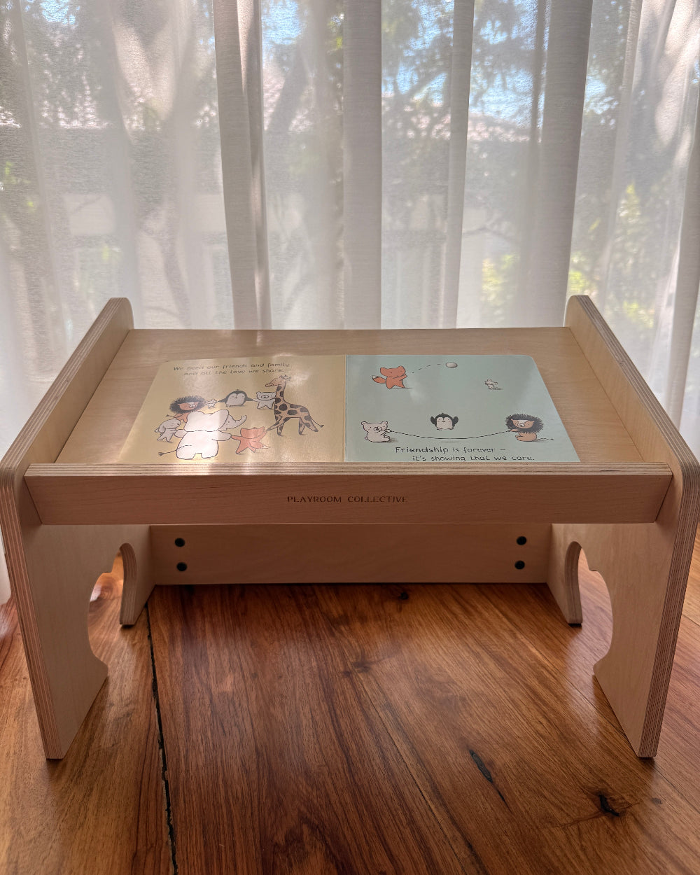 A light natural wood folding table with a glass top, featuring two colorful children's books placed on it, set on a wooden floor in front of sheer white curtains.