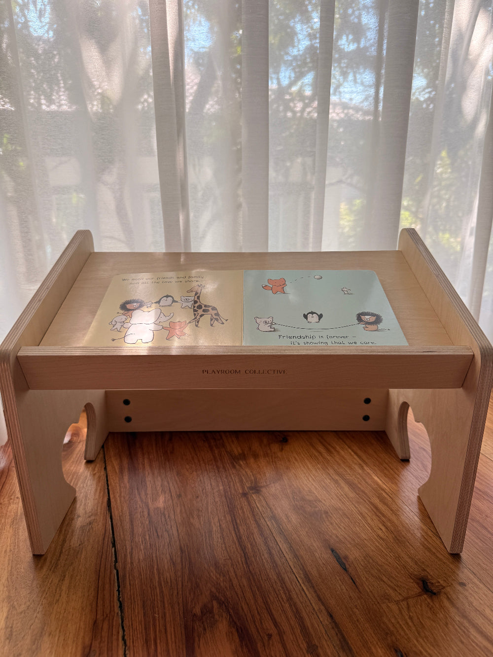 A light natural wood folding table with a glass top, featuring two colorful children's books placed on it, set on a wooden floor in front of sheer white curtains.