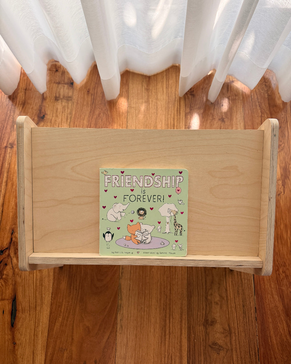 A light natural wood toy shelf or bookcase placed on a hardwood floor in front of white sheer curtains. A square illustrated book or card titled "Friendship is Forever!" rests on the shelf, showing pastel artwork with animals and hearts.