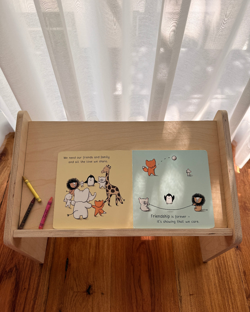 A small wooden children's book stand on a wooden floor in front of sheer white curtains. The stand holds an open children's book with two illustrated pages and two small colored pencils.
