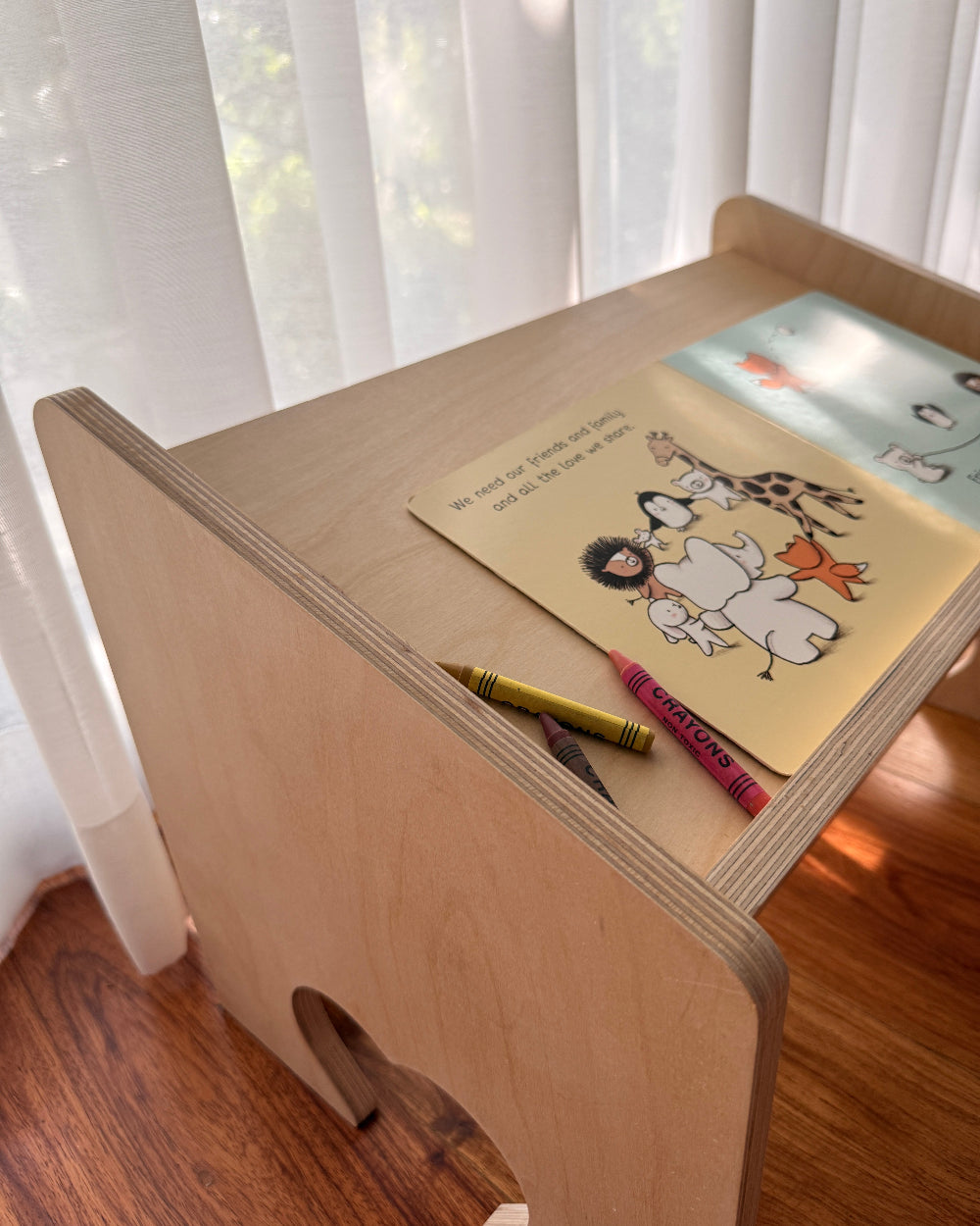 A light natural wood children's desk with a rounded corner and a small cutout, styled with a children's book and crayons on top, set in a room with sheer white curtains and a wooden floor.