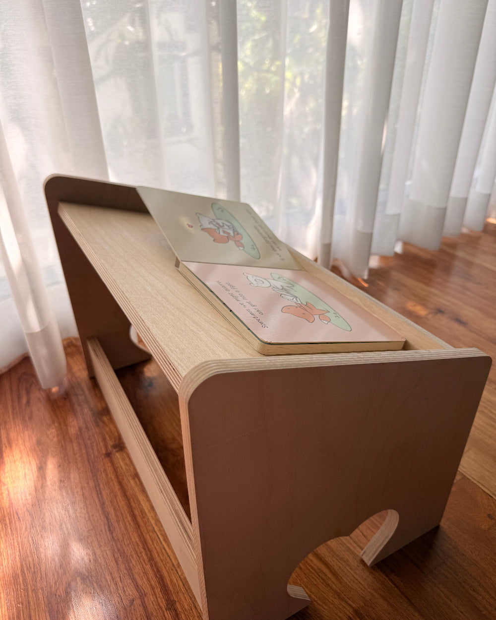 A light natural wood folding desk or lap desk with a slanted top, shown on a hardwood floor in front of sheer white curtains.