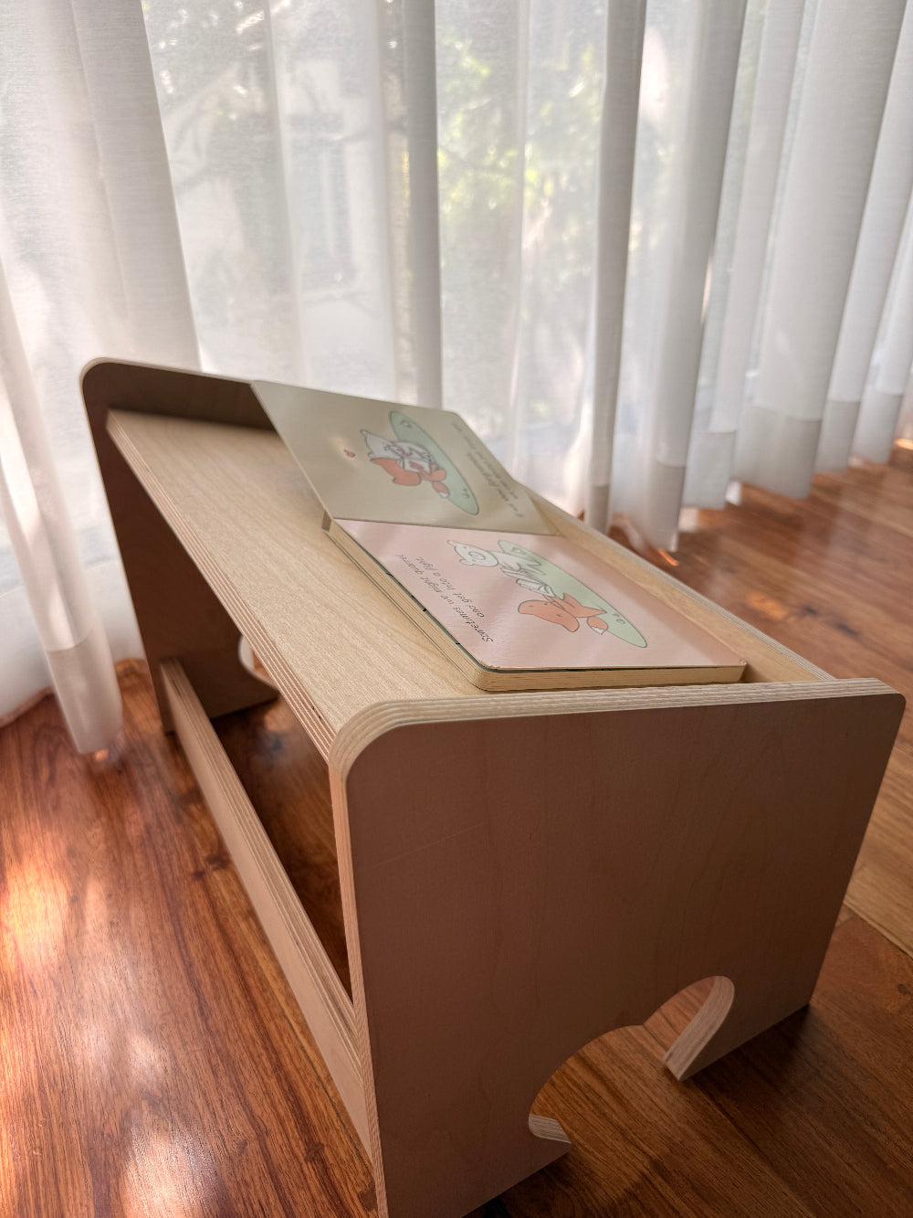 A light natural wood folding desk or lap desk with a slanted top, shown on a hardwood floor in front of sheer white curtains.