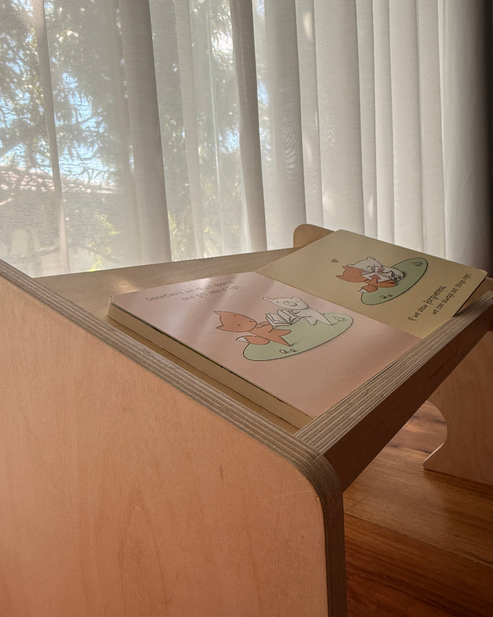A children's picture book with illustrated pages is propped open on a light wood bookshelf or desk next to window blinds.