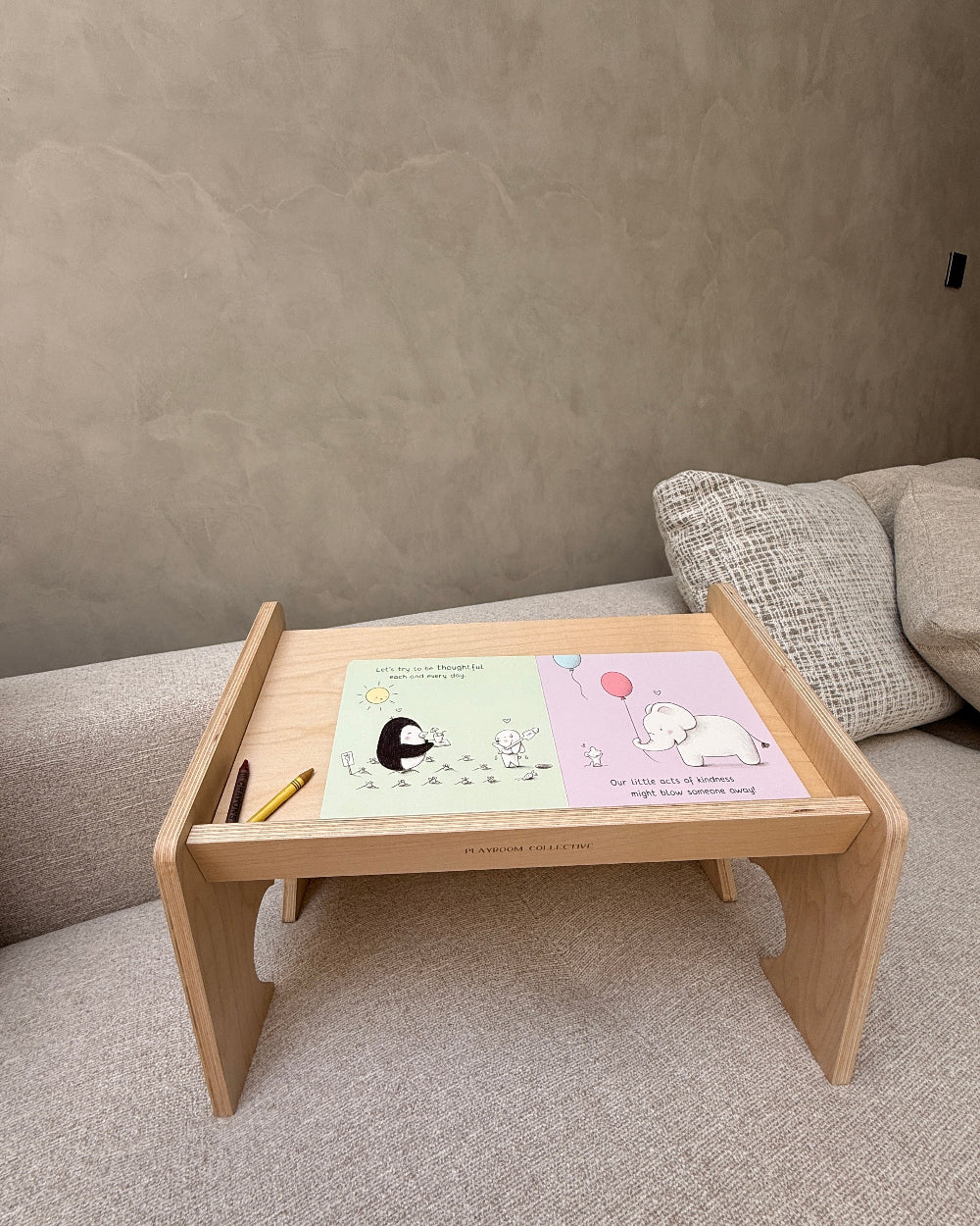 A light natural wood folding lap desk or tray table set up on a textured gray sofa. The desk holds a pastel-colored children's activity book with cartoon illustrations and a small yellow pencil.