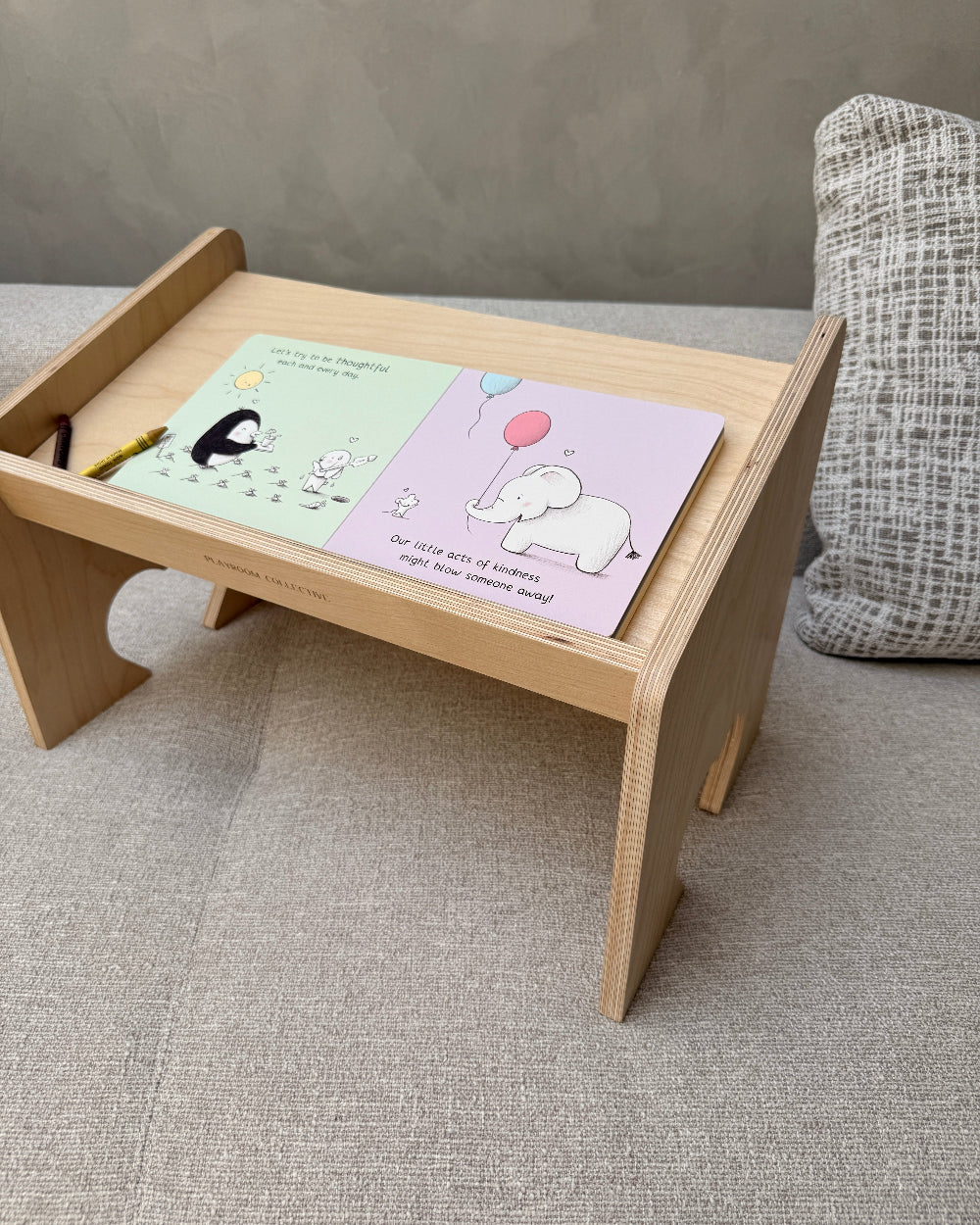 A light natural wood toddler lap desk with a slanted writing tray, shown on a textured gray sofa. The tray holds a pastel coloring book with a penguin and an elephant illustration, along with a small yellow pencil.