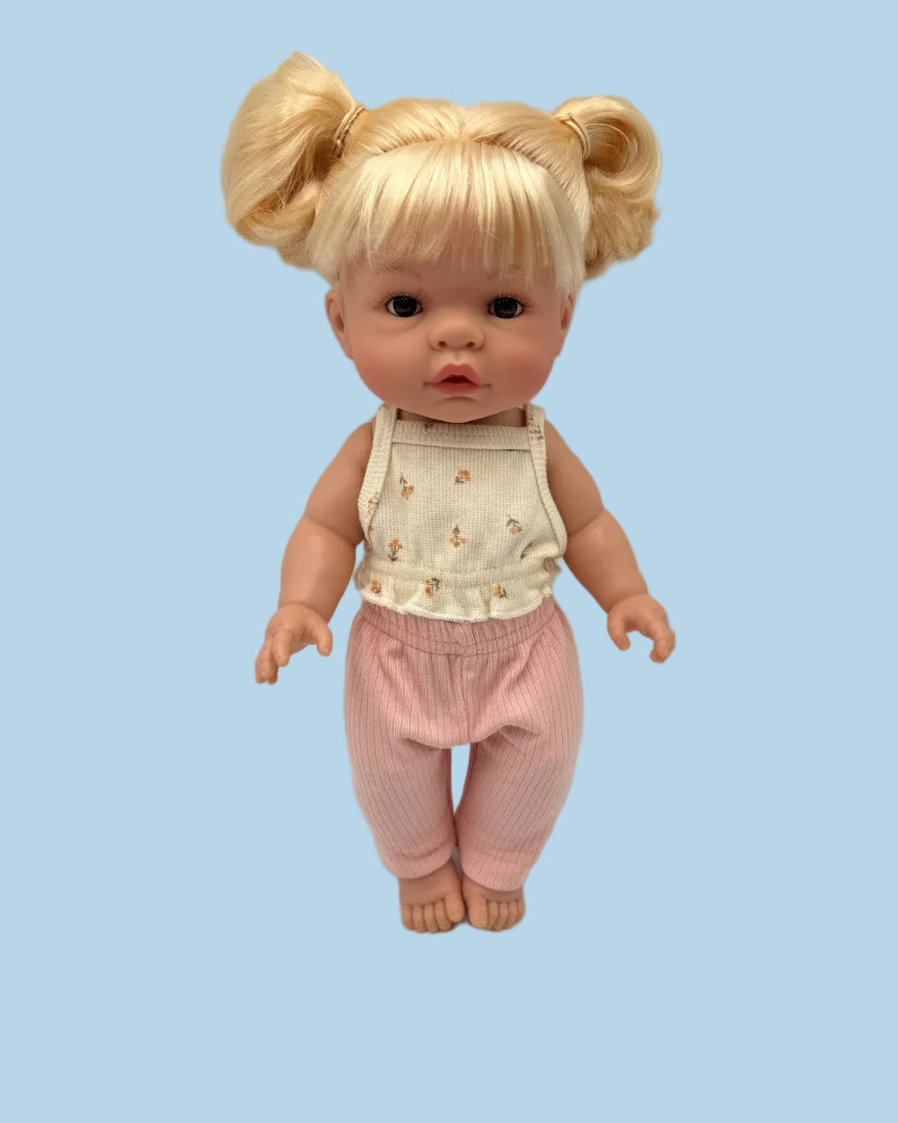 A baby doll with blonde hair in pigtails, wearing a cream sleeveless top with small yellow floral prints and pink pants, standing against a light gray background.