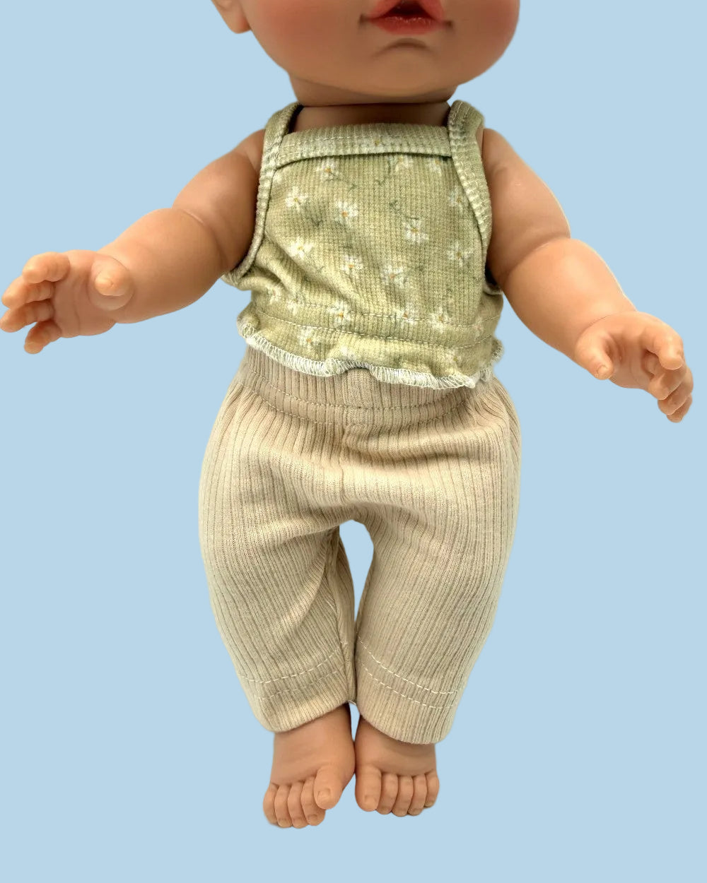 A doll or toy baby wearing a sleeveless light sage green top with a small white floral pattern and ribbed beige pants, posed against a plain white background.
