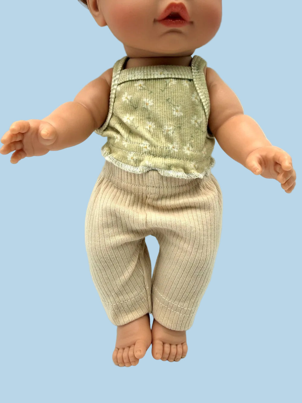 A doll or toy baby wearing a sleeveless light sage green top with a small white floral pattern and ribbed beige pants, posed against a plain white background.