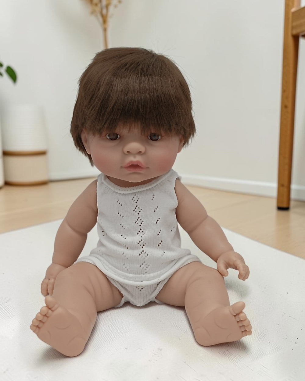 Realistic baby doll with brown hair wearing a white romper on a light surface.