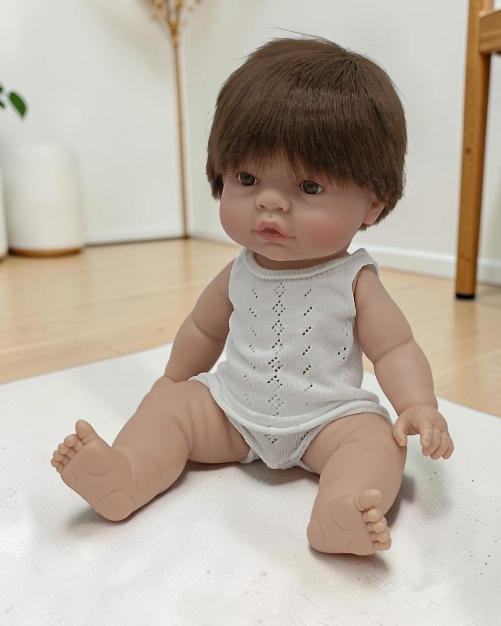 Realistic baby doll with brown hair wearing a white sleeveless top and diaper on a light surface.