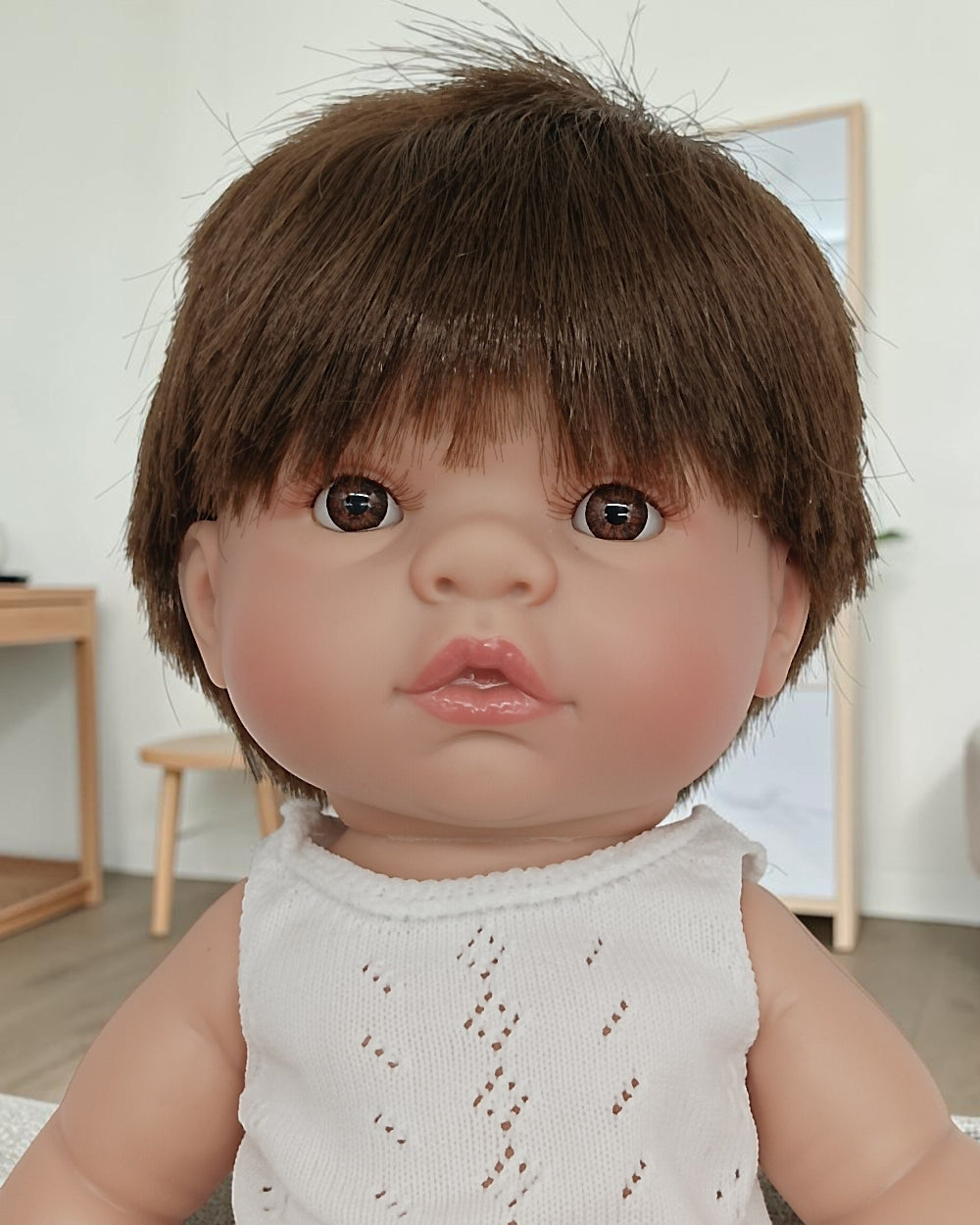 Close-up of a doll with brown hair and a white sleeveless top in a room setting.