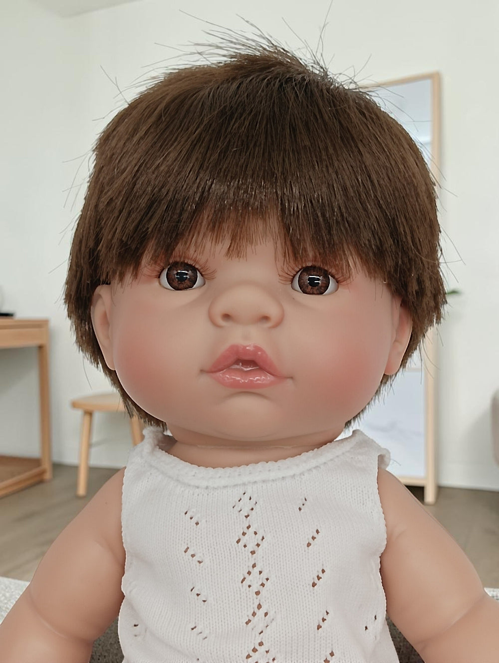 Close-up of a doll with brown hair and a white sleeveless top in a room setting.