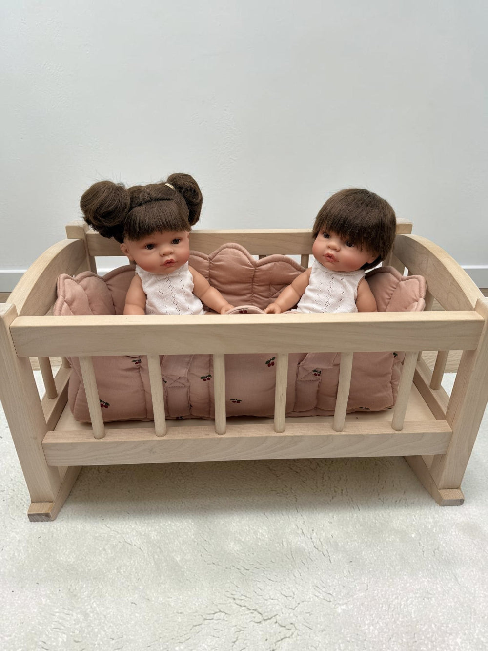 A small wooden doll crib with pink cushioning, holding two baby dolls dressed in white outfits, set against a plain light gray wall and floor.