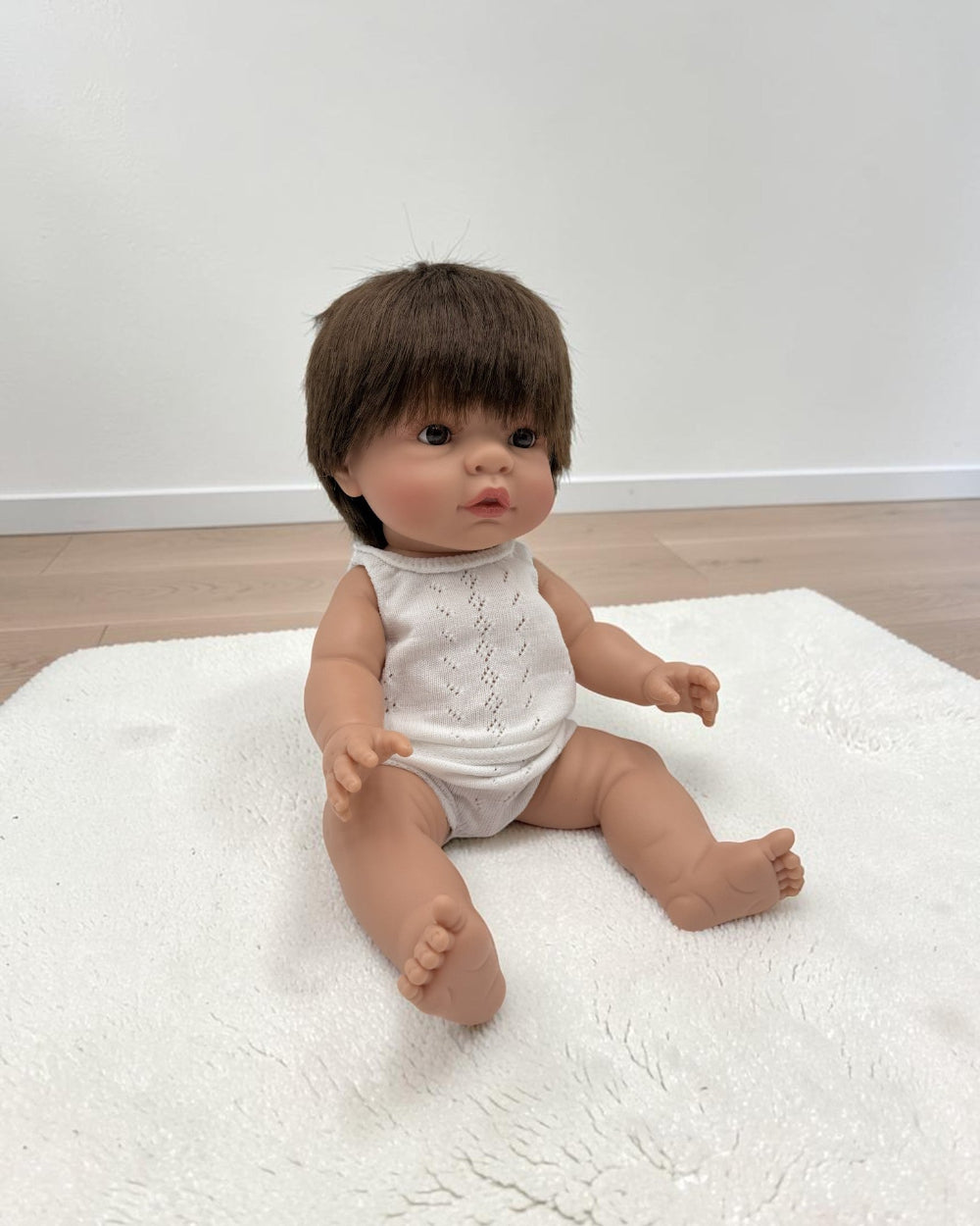 A realistic baby doll with brown hair and skin sits on a white textured rug in a room with light wood flooring and a white wall.