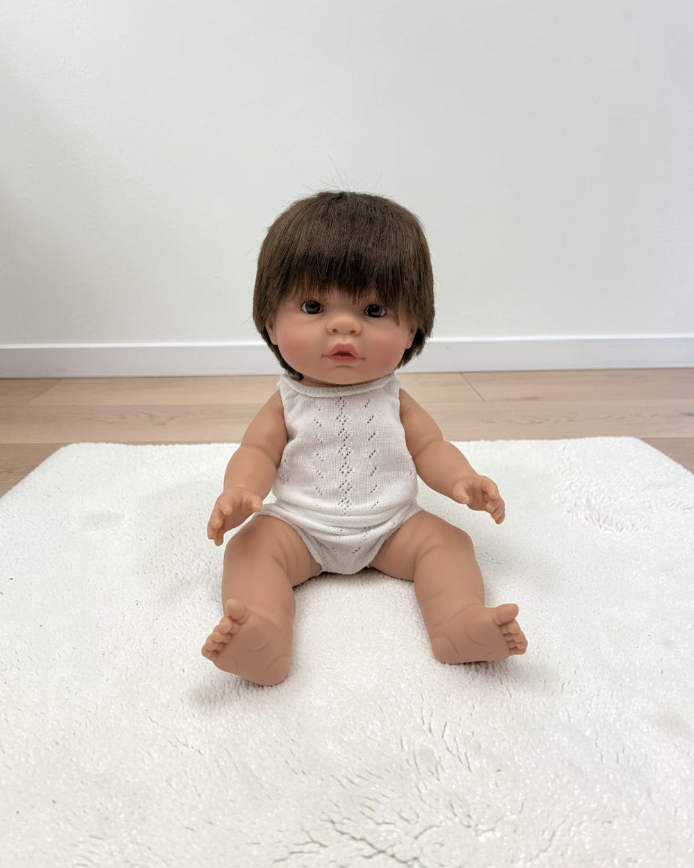 A realistic baby doll with short brown hair sits on a white textured rug in a room with light wood flooring and a white wall.