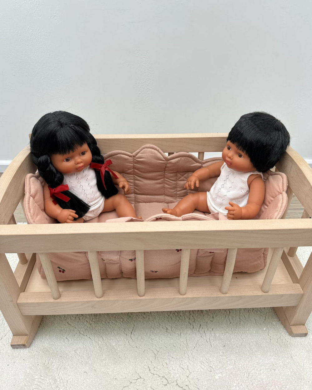 A wooden baby crib with a pink cushion, holding two baby dolls with black hair, one wearing a red and white outfit and the other in a white outfit, set against a plain light gray wall and concrete floor.