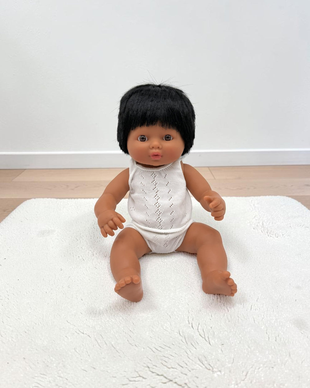 A baby doll with black hair and brown skin sitting on a white textured rug against a white wall and light wood floor.