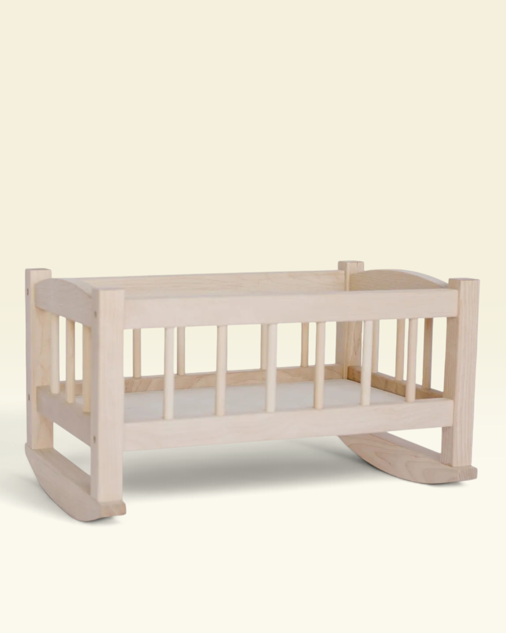 A light natural wood baby rocking cradle with vertical slats and a white mattress, shown against a solid pale cream background.