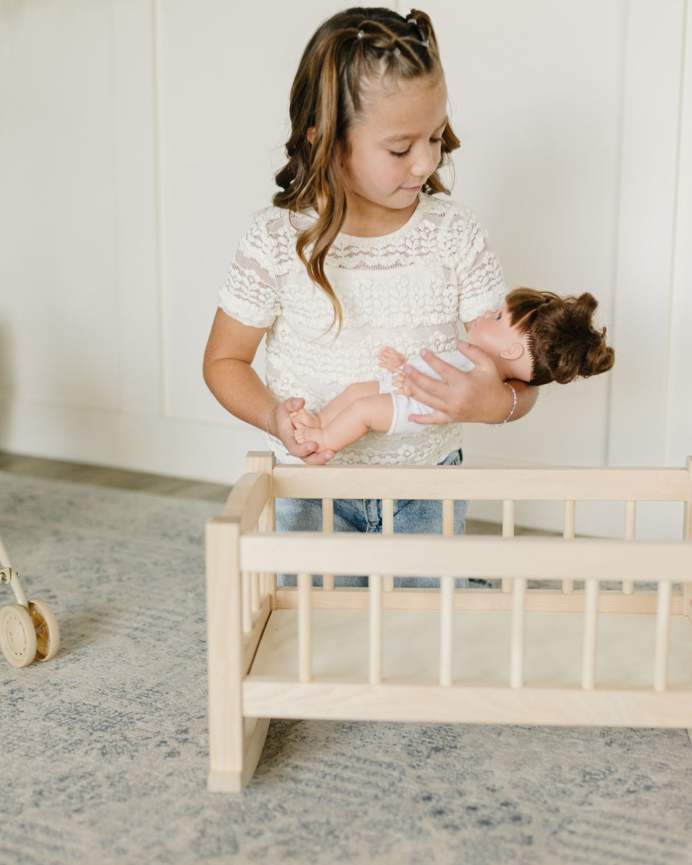 A young girl with braided hair wearing a white lace top and blue jeans holds a doll next to a small light wood crib in a bright room with a patterned rug.