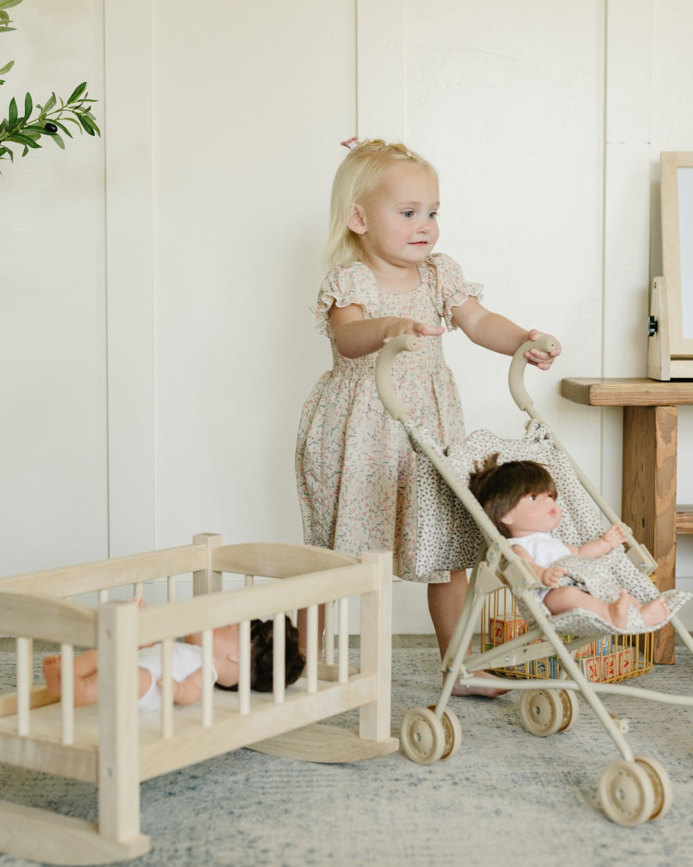 A young toddler girl in a beige patterned dress pushes a light wood toy stroller with a doll inside, next to a wooden rocking cradle with another doll, in a bright, neutral room.