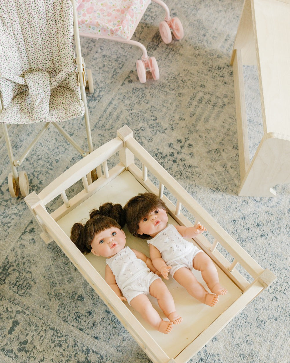 Two baby dolls lying on a cream fabric mattress inside a light wood bassinet, placed on a blue-gray patterned rug in a nursery setting with a stroller and changing table visible in the background.