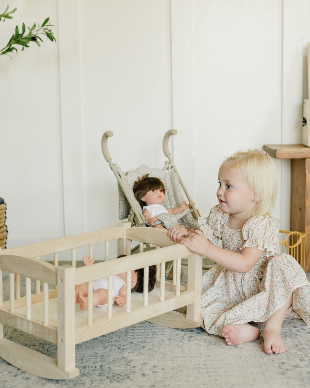 A young blonde toddler in a cream floral dress sits on a gray rug next to a light wood doll crib, playing with a doll in a matching woven rocking chair. The scene is set in a bright, neutral nursery with white paneled walls.