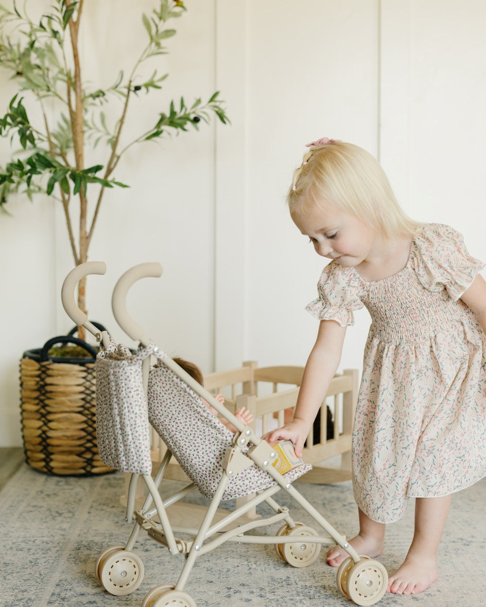 A young blonde girl in a light floral dress pushes a wooden toy stroller with a patterned fabric canopy in a bright, neutral room.