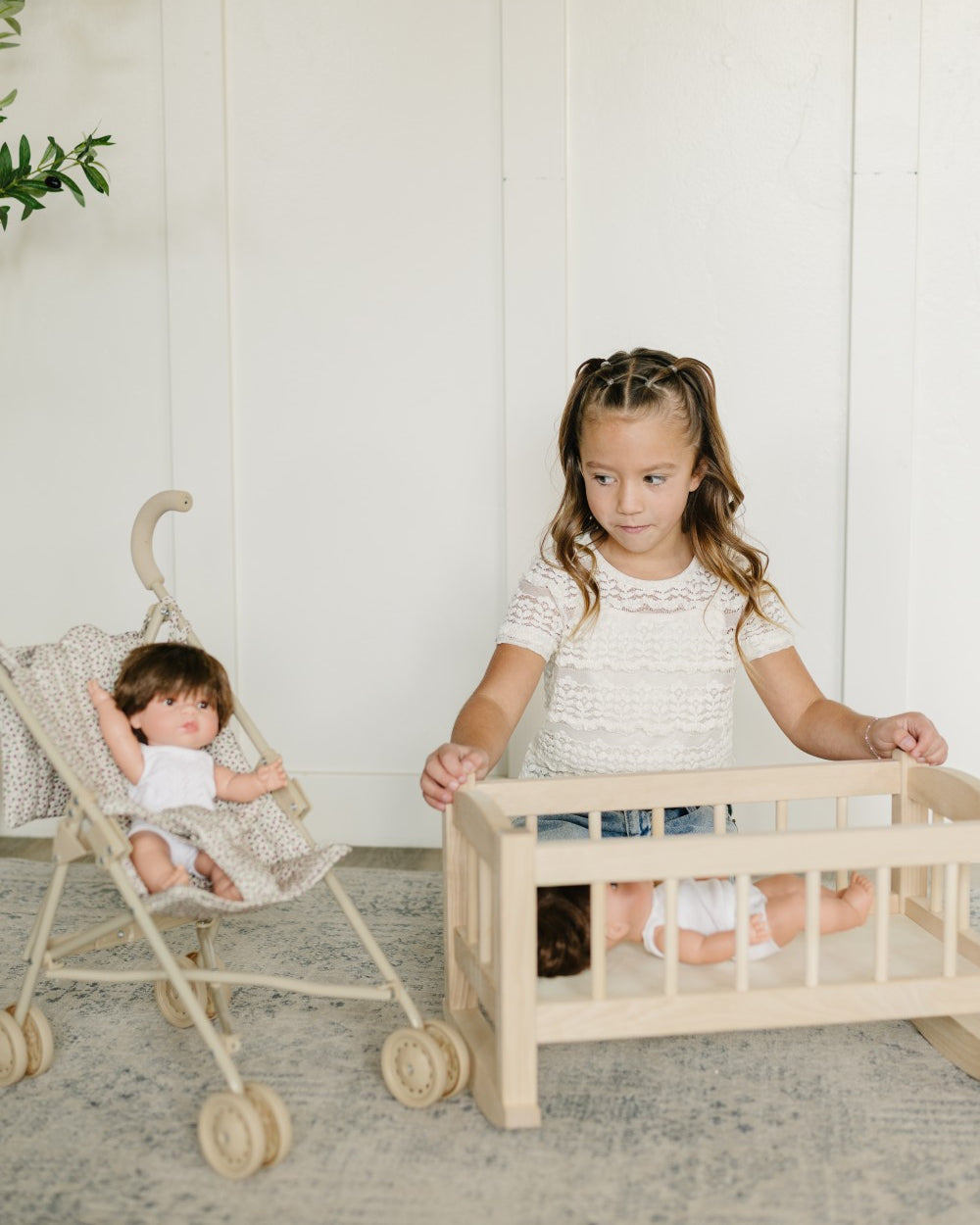 A young girl stands behind a light wood doll crib while a baby doll sits inside, with a stroller holding another doll to the left, set against a white paneled wall and a textured rug.
