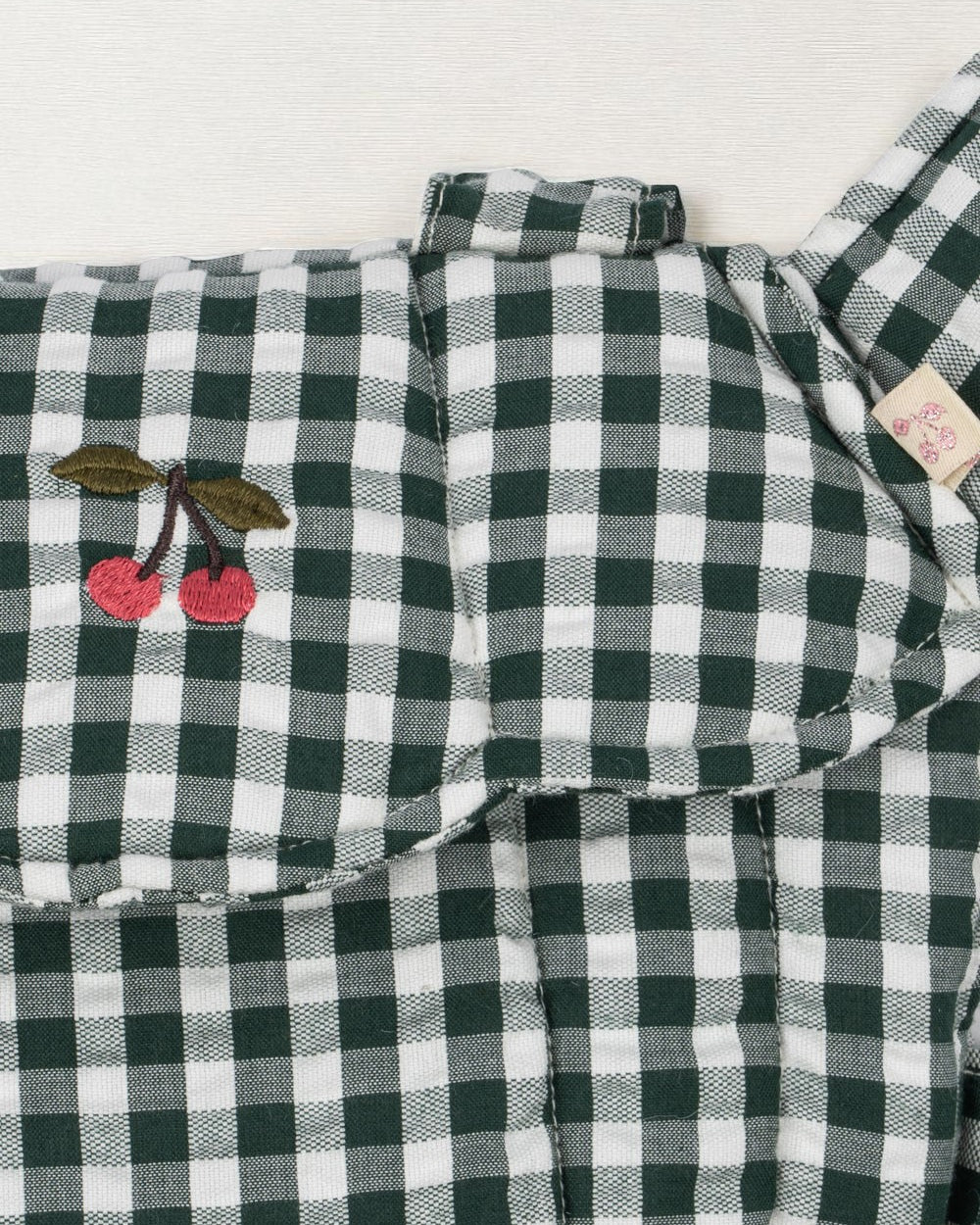 Close-up of embroidered cherry accent on green and white checkered doll carrier showing whimsical farmhouse detail