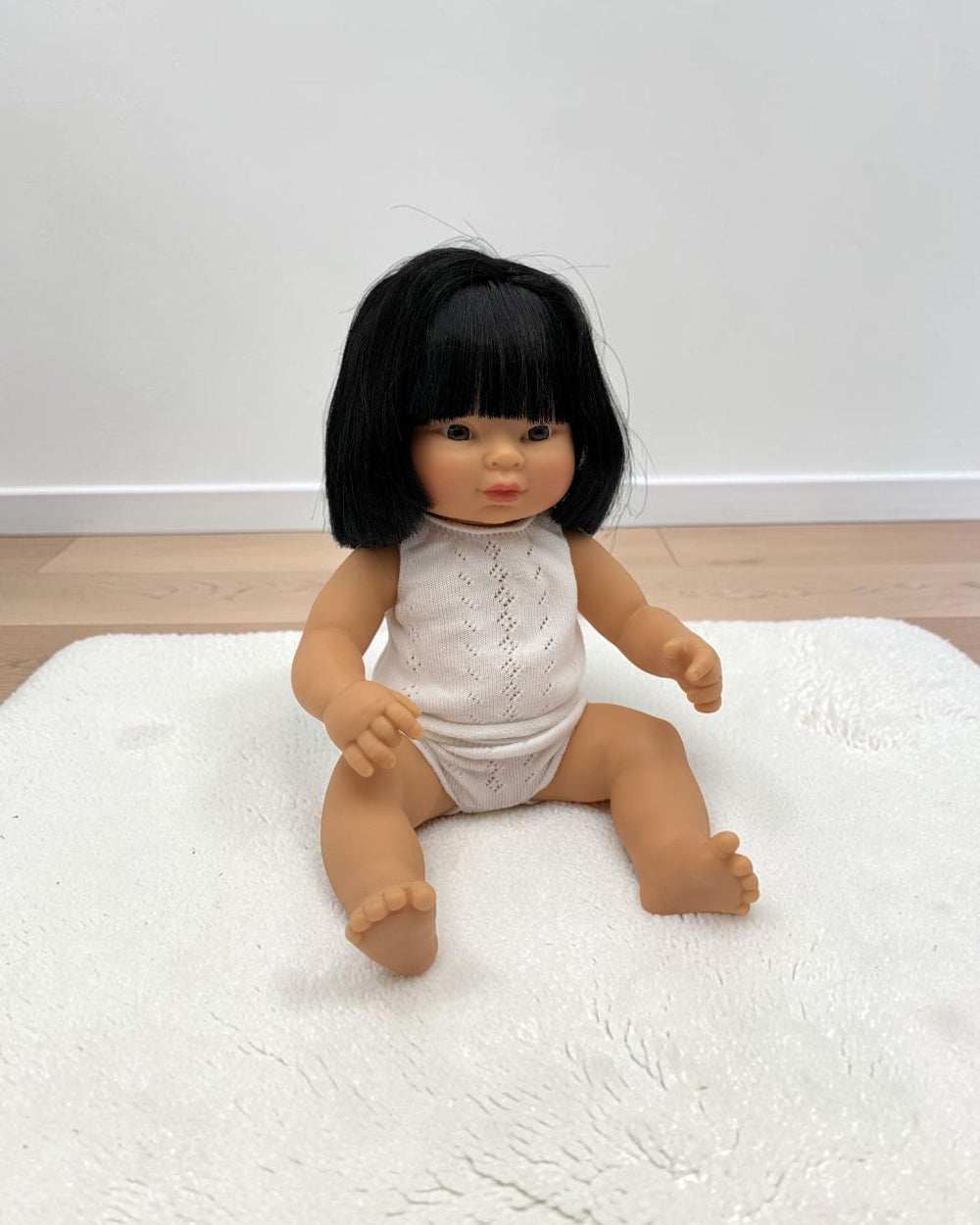 A baby doll with black hair sitting on a white textured rug against a plain white wall and light wood floor.