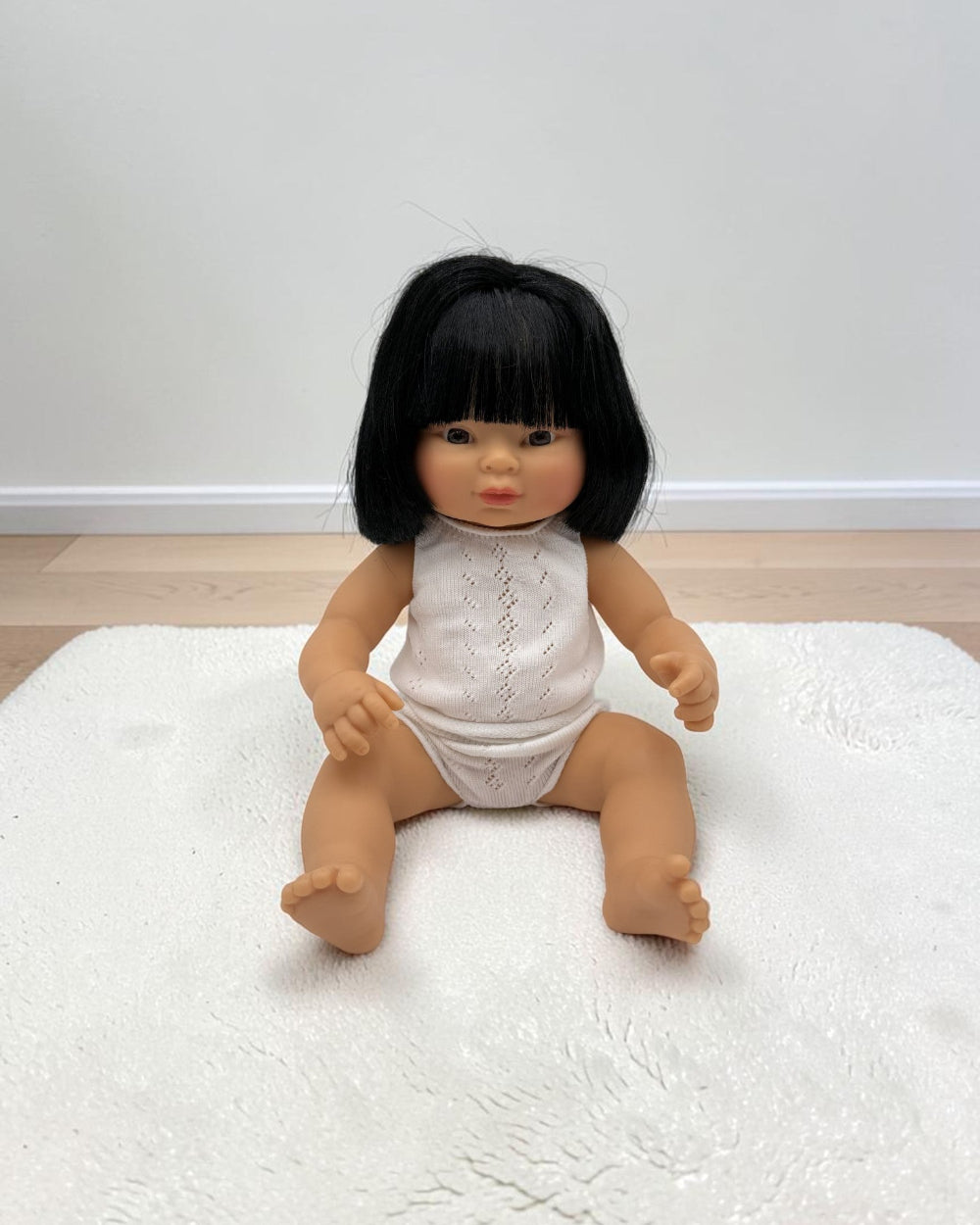A realistic baby doll with black hair sitting on a white textured rug in front of a plain wall.