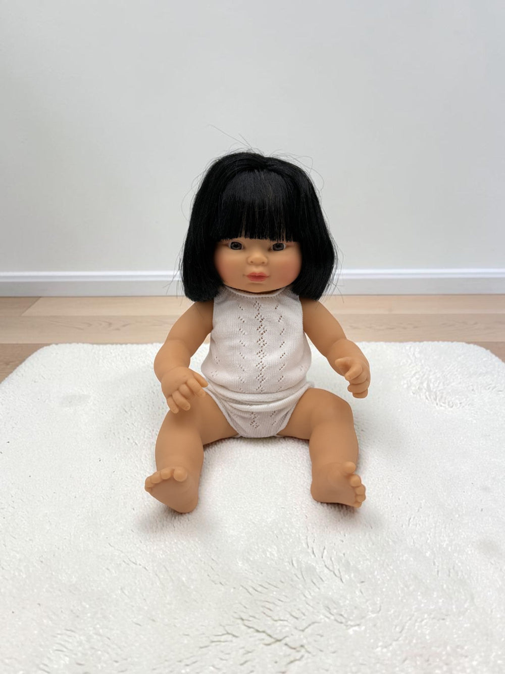 A realistic baby doll with black hair sitting on a white textured rug in front of a plain wall.